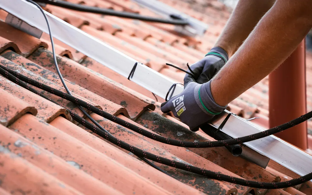 Gloved hands securing solar cables to a mounting rail on a clay-tile roof
